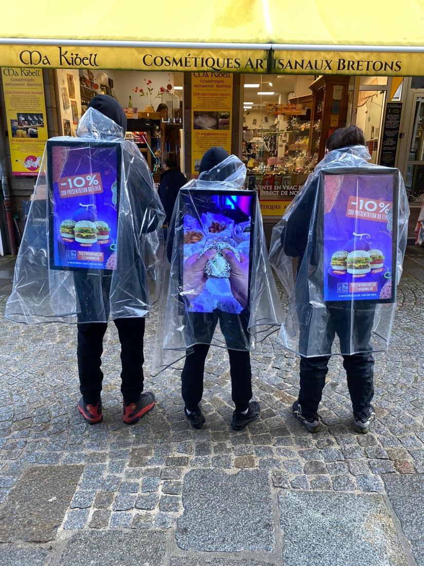 Three promoters in rain ponchos carrying backpack screens outside a shop